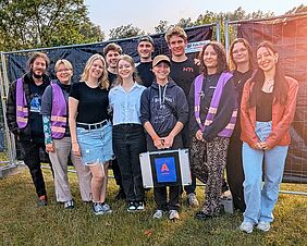 Elf Personen stehen vor einem schwarz verkleideten Bauzaun. Einige von ihnen tragen lilafarbene Westen. Die Person vorne in der Mitte hält einen silbernen Metallkoffer mit einem lilafarbenen "A" für Awareness als Aufschrift auf blauem Grund.
