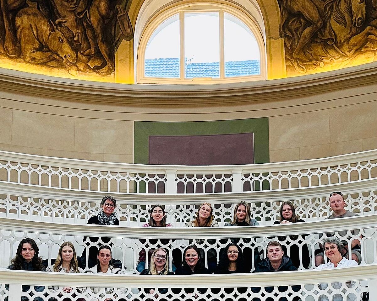 Vierzehn Personen sitzen in einer reichlich verzierten mehrstöckigen Empore. In der mittleren Etage sitzen sechs Personen, in der unteren Etage sitzen acht Personen. Alle lächeln freundlich in die Kamera.