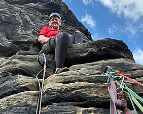Zu sehen ist eine Frau in Klettermontur und rotem T-Shirt. Sie lächelt der Kamera von einem hohen Felsen entgegen. Im Hintergrund hellblauer Himmel und weiße Wolken
