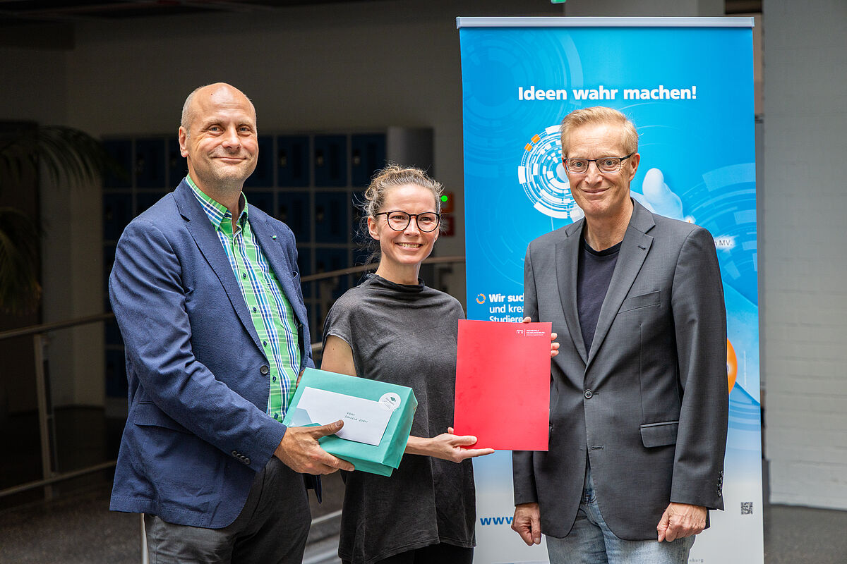 Drei förmlich angezogene Personen lächeln freundlich in die Kamera. Im Hintergrund ist ein blauer Rollup-Banner mit der Aufschrift „Ideen werden wahr“ zu erkennen. Die Frau in der Mitte trägt eine Brille und zugebundene Haare. Sie hält eine rote Mappe hoch, einer der Männer neben ihr hält ein türkisfarbenes Geschenkpaket mit einem weißen Umschlag darauf, ebenfalls sichtbar in die Kamera.