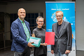 Drei förmlich angezogene Personen lächeln freundlich in die Kamera. Im Hintergrund ist ein blauer Rollup-Banner mit der Aufschrift „Ideen werden wahr“ zu erkennen. Die Frau in der Mitte trägt eine Brille und zugebundene Haare. Sie hält eine rote Mappe hoch, einer der Männer neben ihr hält ein türkisfarbenes Geschenkpaket mit einem weißen Umschlag darauf, ebenfalls sichtbar in die Kamera.