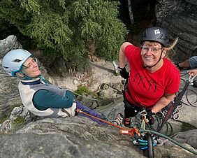 Zu sehen sind zwei Frauen in Klettermontur am Felsen. Eine lehnt entspannt am Stein, die andere schaut gen Kamera, die sich über den Köpfen der Personen befindet. 