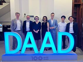 Sieben Personen stehen hinter einer großen Lichtinstellation, die aus blau leuchtenden Buchstaben bestehen, die die Abkürzung DAAD formen. Die Personen sind förmlich, in Kleidern oder Anzügen gekleidet und lächeln freundlich in die Kamera.