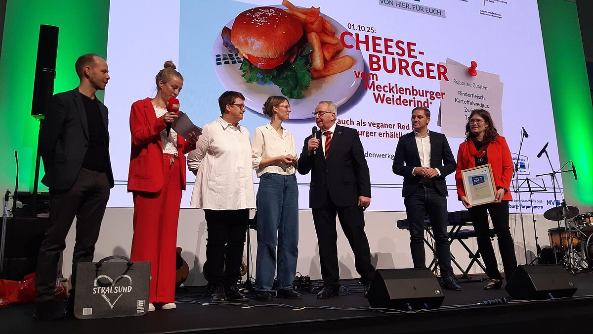 Zu sehen ist eine Preisverleihungssituation. Sieben Personen stehen auf einer Bühne. Die Moderatorin im roten Hosenanzug überprüft ihre Moderationskarten. Eine Frau, die rechts außen steht, hält eine gerahmte Urkunde vor sich. In der Mitte spricht der Landwirtschaftsminister Till Backhaus in ein Mikrofon. Hinter den Personen ist ein Cheeseburger auf einer Leinwand eingeblendet. 