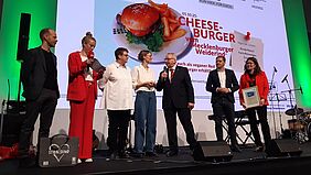Zu sehen ist eine Preisverleihungssituation. Sieben Personen stehen auf einer Bühne. Die Moderatorin im roten Hosenanzug überprüft ihre Moderationskarten. Eine Frau, die rechts außen steht, hält eine gerahmte Urkunde vor sich. In der Mitte spricht der Landwirtschaftsminister Till Backhaus in ein Mikrofon. Hinter den Personen ist ein Cheeseburger auf einer Leinwand eingeblendet.