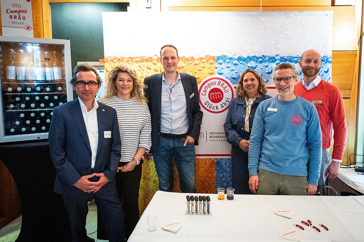 Sechs Personen haben sich zum gemeinsamen Gruppenfoto aufgestellt und lächeln freundlich in die Kamera. Im Hintergrund befindet sich ein Aufsteller, auf dem das Logo vom Campus Bräu zu erkennen ist. 