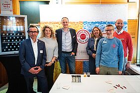 Sechs Personen haben sich zum gemeinsamen Gruppenfoto aufgestellt und lächeln freundlich in die Kamera. Im Hintergrund befindet sich ein Aufsteller, auf dem das Logo vom Campus Bräu zu erkennen ist. 