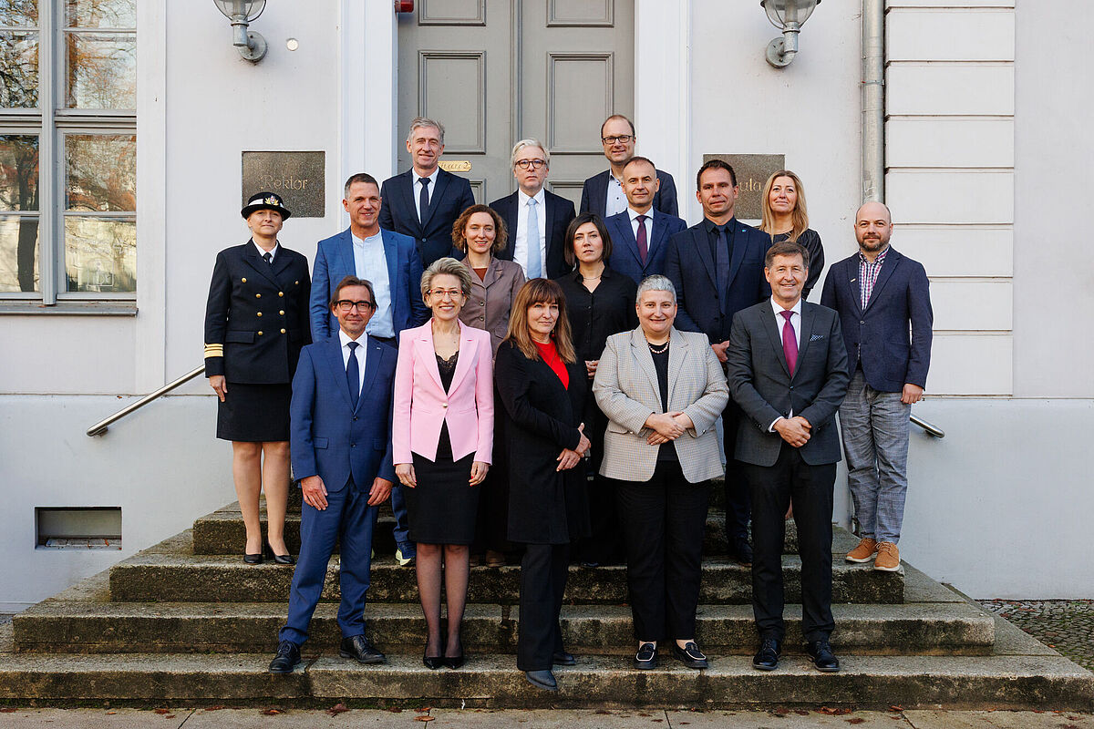 16 Personen stehen in drei Reihen auf einer Treppe und lächeln geübt und freundlich in die Kamera. Im Hintergrund befindet sich ein weißes Haus mit einer großen grauen Tür. Die Personen auf der Treppe sind amtlich in Anzügen und festlichen Zweiteilern gekleidet. 