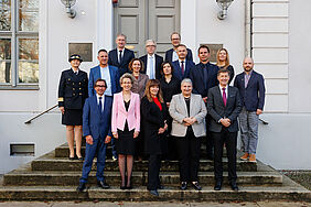 16 Personen stehen in drei Reihen auf einer Treppe und lächeln geübt und freundlich in die Kamera. Im Hintergrund befindet sich ein weißes Haus mit einer großen grauen Tür. Die Personen auf der Treppe sind amtlich in Anzügen und festlichen Zweiteilern gekleidet. 
