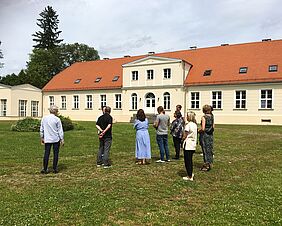 Mehrere Personen in sommerlicher Kleidung stehen auf einer grünen Wiese. Im Hintergrund befindet sich ein helles, großes Gutshaus.