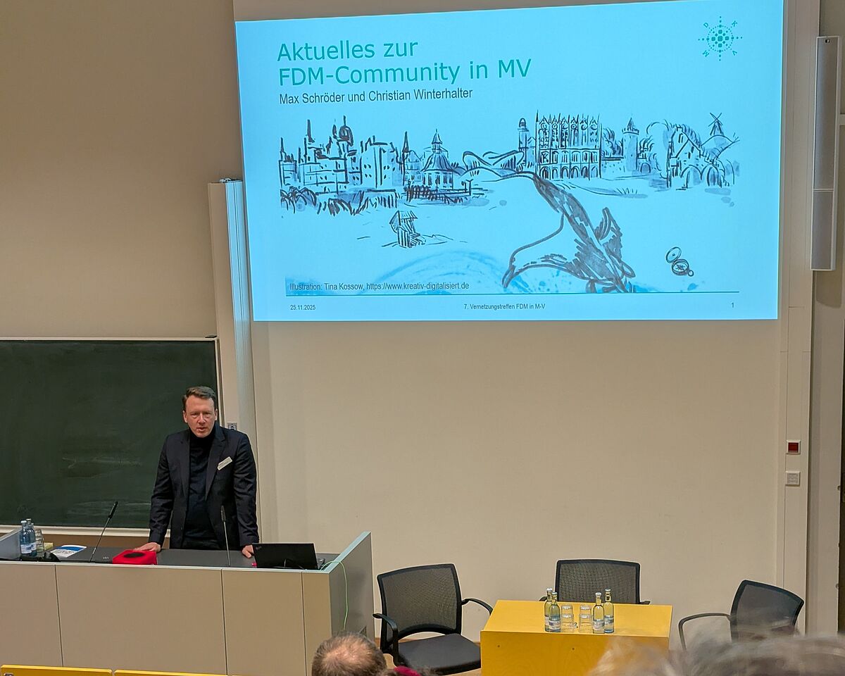 Ein Mann steht hinter einem Podium, vor einer grünen Tafel. Neben der Tafel wird eine Präsentation auf weißer Wand eingeblendet, die den Titel „Aktuelles zur FDM-Community in MV“ trägt. Der Redner trägt ein dunkles Sakko, darunter einen dunklen Rollkragenpullover.