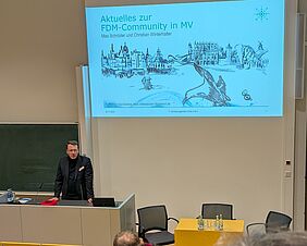 Ein Mann steht hinter einem Podium, vor einer grünen Tafel. Neben der Tafel wird eine Präsentation auf weißer Wand eingeblendet, die den Titel „Aktuelles zur FDM-Community in MV“ trägt. Der Redner trägt ein dunkles Sakko, darunter einen dunklen Rollkragenpullover.