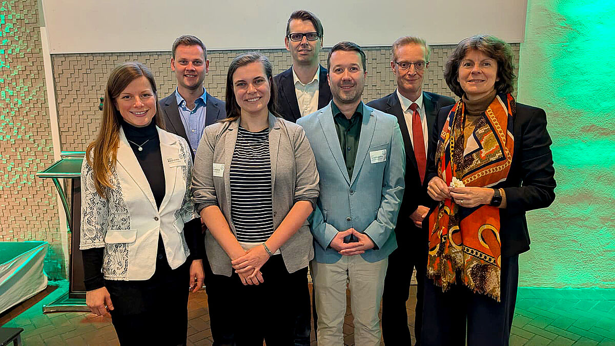 Auf dem Foto sind sieben Personen in zwei Reihen aufgestellt. Im Hintergrund befindet sich eine grün beleuchtete Wand mit verschiedenen Strukturen. Die Personen sind festlich bis förmlich gekleidet und lächeln freundlich in die Kamera.