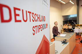 Im Vordergrund befindet sich ein großer Aufsteller mit der Aufschrift „Deutschlandstipendium“. Im Hintergrund spricht ein junger Mann an einem Podium.