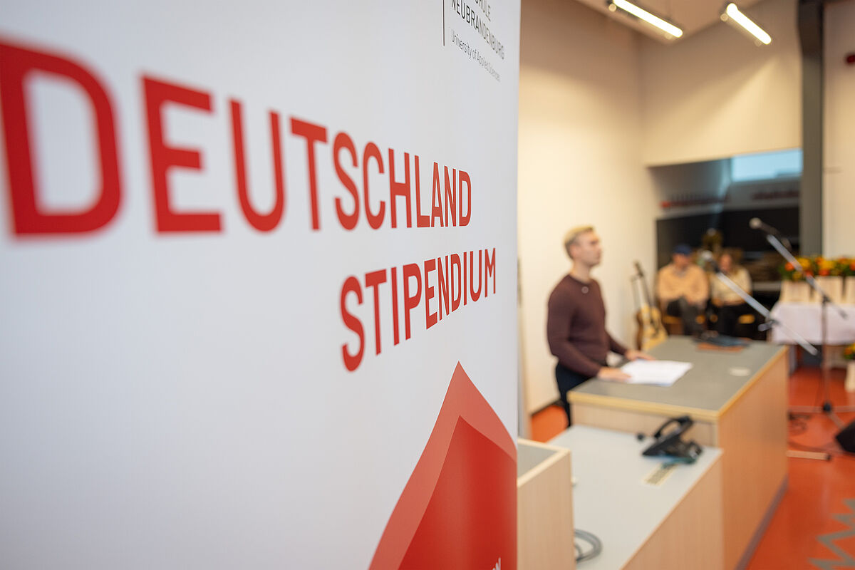 Im Vordergrund befindet sich ein großer Aufsteller mit der Aufschrift „Deutschlandstipendium“. Im Hintergrund spricht ein junger Mann an einem Podium.