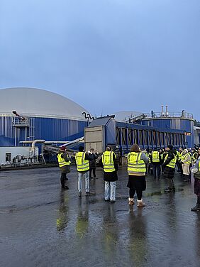 Personen mit gelben Warnwesten stehen vor einer Biogasanlage. Einige Personen filmen und fotografieren mit ihren Smartphones.