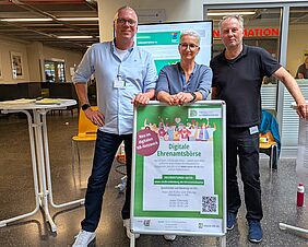 Auf dem Foto sind drei Personen zu erkennen, die freundlich in die Kamera schauen. Vor ihnen steht ein Aufsteller mit einem Hinweis auf die Digitale Ehrenamtsbörse der Stadt Neubrandenburg.