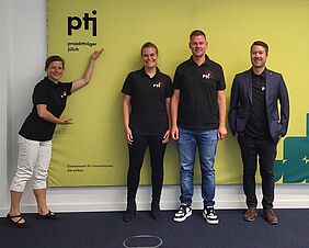 Auf dem Foto befinden sich vier Personen vor einer grünen Wand mit der Aufschrift „Projektträger Jülich“. Die Frau ganz links posiert mit den Händen in Richtung der Aufschrift, die anderen drei Personen lächeln freundlich in die Kamera. Sie alle tragen schwarze Oberteile mit dem Logo von Plasma Grain Innovation.