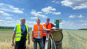 Auf diesem Gruppenfoto sind drei Männer in orangefarbenen Warnwesten zu erkennen. Sie stehen mit einem Entfernungsmesser auf einem weiten Feld. Die Szenerie ist freundlich und wirkt warm und sonnig.