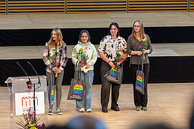 Vier Jugendliche stehen gespannt auf einer hellen Bühne. Sie halten Blumen und farbige Jutebeutel mit Hochschullogo in den Händen. Bei den Personen handelt es sich um Juniorstudierende.