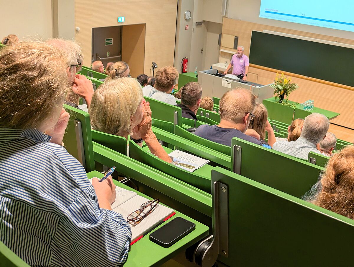Personen sitzen in einem Hörsaal. Unscharf im Vordergrund ist ein Referent zu erkennen, der an einem Pult vor einer Tafel steht. Die Stühle des Hörsaals sind treppenförmig angeordnet. Das Motiv ist aus der Perspektive eines Gastes in der hintersten, obersten Reihe fotografiert.
