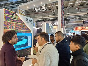 Das Bild zeigt eine typische Szene einer großen Messe. An einem Informationsstand unterhalten sich Aussteller und Besucher, im Hintergrund sind weitere Stände und Menschen in den Gängen zu sehen. An der Decke der Messe befinden sich lange Neonröhren, Metallelemente und massiver Beton.