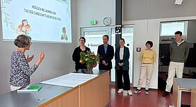 Die fünf Vortragenden der Veranstaltung stehen aufgereiht im Hörsaal nebeneinander. Alle schauen in Richtung Rednerpult, wo eine weitere Person zu ihnen spricht. An die Wand im Hintergrund wird eine Präsentation projiziert, auf der „Herzlich willkommen zum Tag der Landschaftsarchitektur 2026“ zu lesen ist.