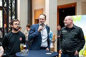 Drei Männer stehen an einem Stehtisch. Einer von ihnen hält ein Mikrofon, in das er spricht. Auf dem Stehtisch befinden sich verschieden gefüllte Biergläser. 