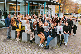 Ein Gruppenfoto mit mehr als 40 Personen. Die Menschen stehen und sitzen vor einem Gebäude und lächeln freundlich in die leicht höher positionierte Kamera. Im Hintergrund befinden sich Autos und herbstlich eingefärbte Bäume.