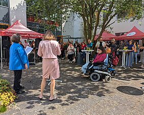 Zwei Frauen sprechen im Innenhof des Hochschulhauses zu Publikum. Es sind rote Pavillons und Stehtische sowie die Gesichter der Gäste zu erkennen. Die Rednerinnen stehen mit dem Rücken zur Kamera. Die links stehende Person trägt einen sattblauen Blazer, die Person neben ihr eine pastellrosafarbene Kombination aus Jacke und Rock.