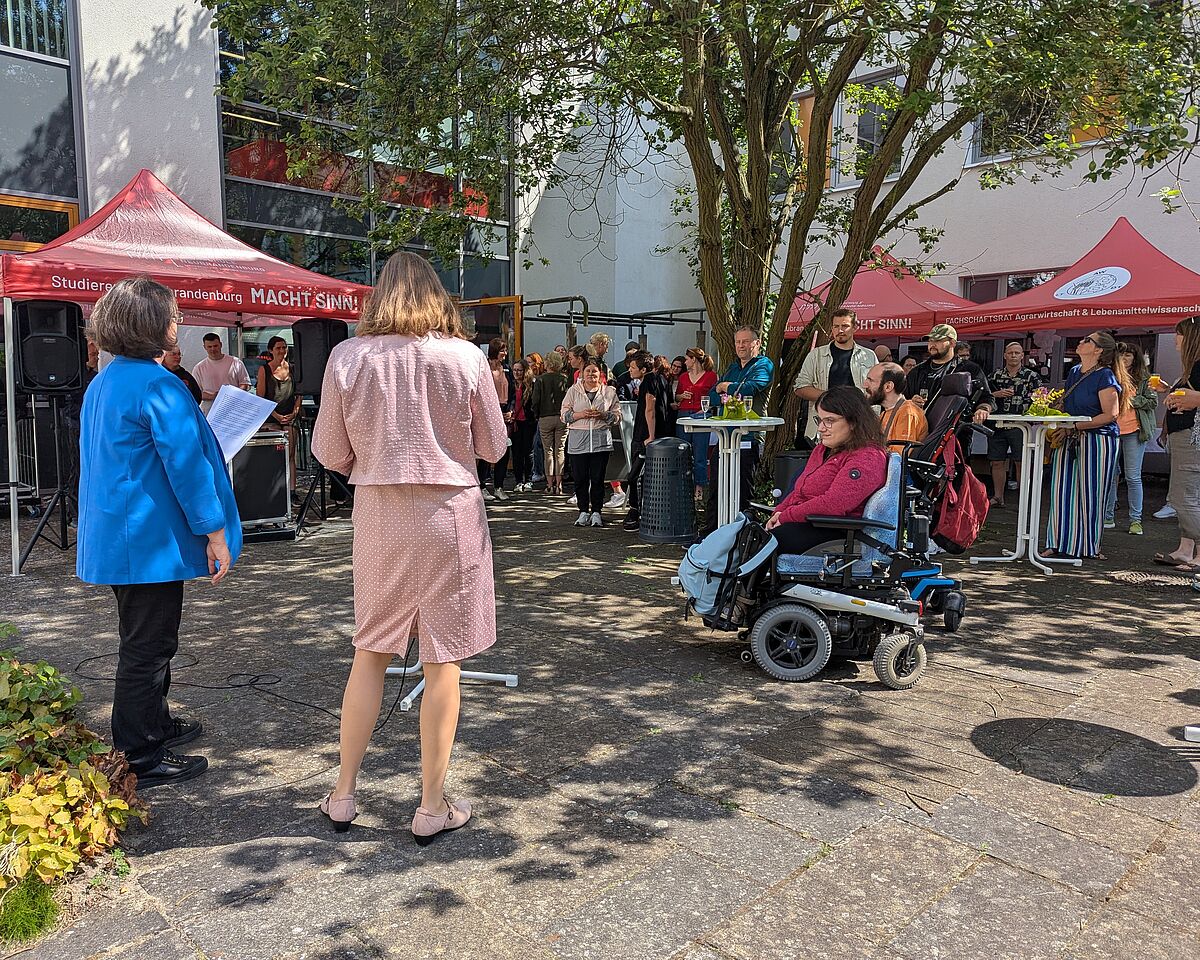 Zwei Frauen sprechen im Innenhof des Hochschulhauses zu Publikum. Es sind rote Pavillons und Stehtische sowie die Gesichter der Gäste zu erkennen. Die Rednerinnen stehen mit dem Rücken zur Kamera. Die links stehende Person trägt einen sattblauen Blazer, die Person neben ihr eine pastellrosafarbene Kombination aus Jacke und Rock.