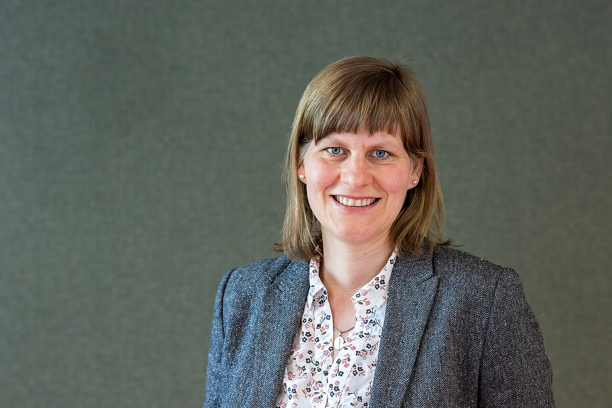 Zu sehen ist eine Frau, die freundlich in die Kamera lächelt. Sie trägt Perlenohrringe und eine helle Bluse mit Blumenmuster, darüber einen Blazer. Im Hintergrund befindet sich unscharf eine Wand.