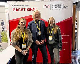 Auf dem Foto stehen die Agrarbotschafterinnen Amy Wolters und Johanna Emmler auf der Deutschen Lohnunternehmer-Tagung mit Prof. Dr. Rainer Langosch. Im Hintergrun befindet sich ein Messeaufsteller mit dem Logo der Hochschule Neubrandenburg. Alle Personen lächeln freundlich in die Kamera.