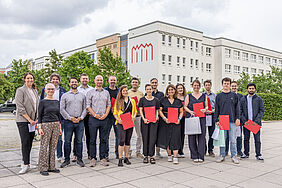 Ein Gruppenfoto. Zu sehen sind unterschiedliche Personen, die freundlich in die Kamera lächeln. Im Hintergrund befindet sich ein Gebäude – das Hauptgebäude der Hochschule