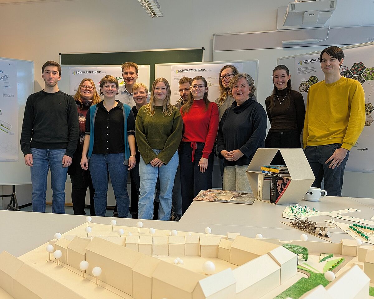 Zwölf Personen lächeln für ein gemeinsames Gruppenfoto in die Kamera. Vor ihnen befindet sich ein landschaftsarchitektonisches Modell, welches Bäume, Häuser und eine Stadtmauer andeutet. Hinter ihnen befinden sich Flipcharts mit Postern, auf denen unter anderem das Wort „Schwarmprinzip“ zu erkennen ist. 