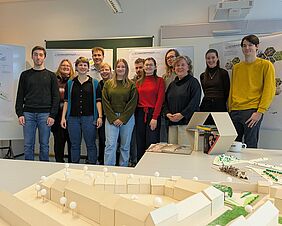 Zwölf Personen lächeln für ein gemeinsames Gruppenfoto in die Kamera. Vor ihnen befindet sich ein landschaftsarchitektonisches Modell, welches Bäume, Häuser und eine Stadtmauer andeutet. Hinter ihnen befinden sich Flipcharts mit Postern, auf denen unter anderem das Wort „Schwarmprinzip“ zu erkennen ist. 