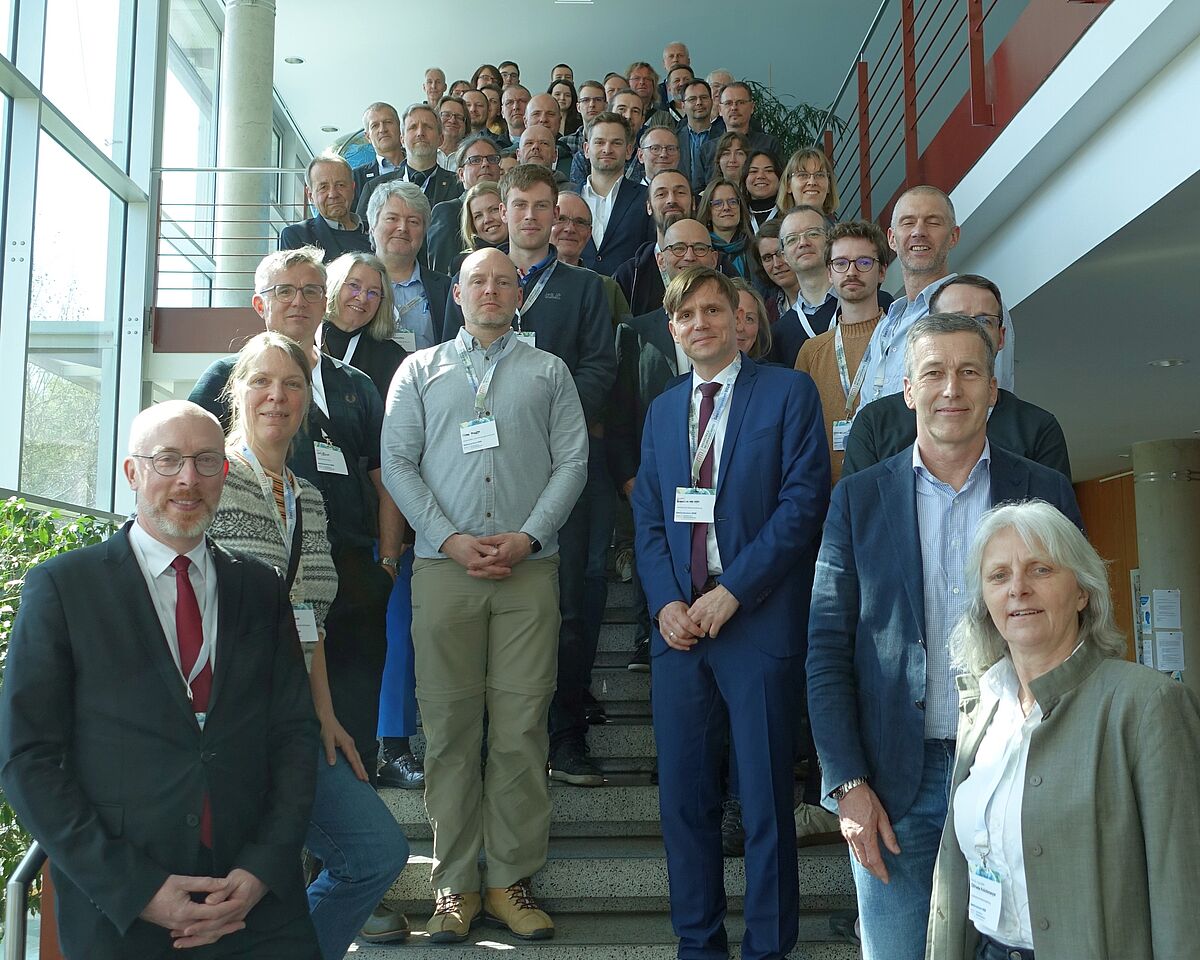 Ein Gruppenfoto. Auf einer hohen Treppe nehmen viele Menschen für ein Gruppenfoto Platz. Sie lächeln freundlich in die Kamera. Die Personen sind vorrangig förmlich gekleidet.