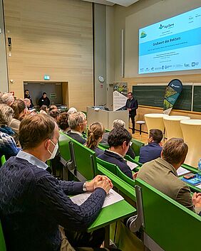 Eine Person spricht an einem Pult in einem Hörsaal vor Publikum. Die Reihen sind gut gefüllt. Neben dem Rednerpult befinden sich drei Stehtische. An der Wand im Hintergrund ist eine Präsentation zum Forschungsprojekt AgriSens zu erkennen.