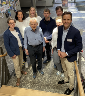 Acht Personen lächeln freundlich in die Kamera. Es handelt sich um eine leichte Draufsicht. Die Kamera wurde auf einer Treppe platziert, die im Foto durch ihr Gelände an den rechten und linken Bildrändern sichtbar wird.