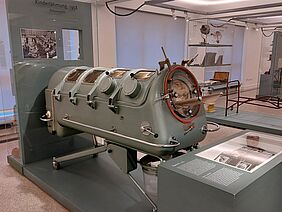 Zu sehen ist ein Ausstellungsraum, in dem sich eine sogenannte Eiserne Lunge befindet. Diese ist körpergroß und erinnert in der Form an ein U-Boot. Schautafeln im Vordergrund scheinen über Funktion und Historie des Geräts aufzuklären.