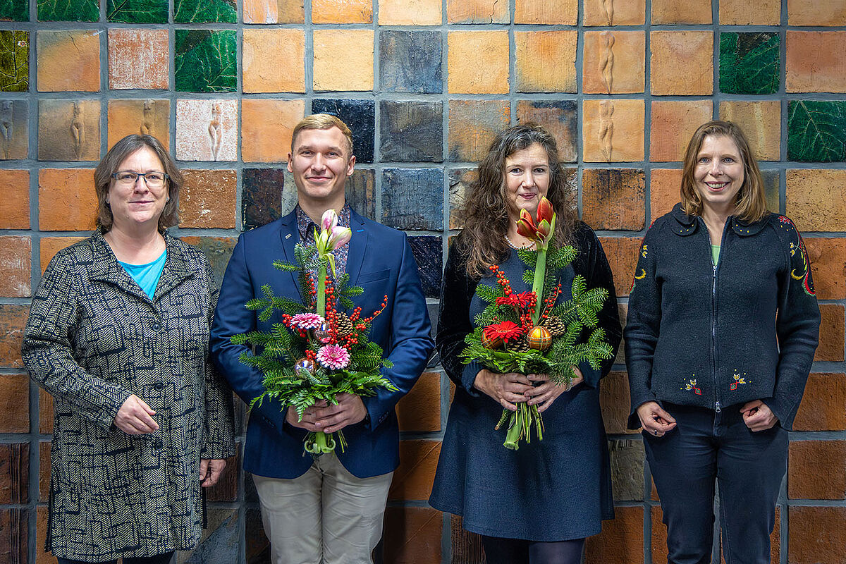Vier Personen lächeln freundlich in die Kamera. Die zwei in der Mitte positionierten Menschen halten jeweils einen festlichen Blumenstrauß in der Hand. Es handelt sich bei den zuletzt genannten um den wiedergewählten Studentischen Prorektor und die wiedergewählte Prorektorin für Studium, Lehre, Weiterbildung und Evaluation. Links und rechts von ihnen stehen die Stellvertretende Vorsitzende sowie die Vorsitzende des Senats. 