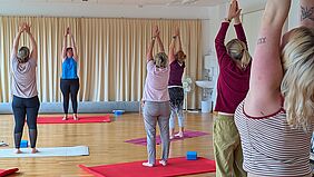 Zu sehen sind sechs Frauen in Yogapositionen. Sie tragen entspannte Kleidung. Eine der Frauen, wahrscheinlich die Yogalehrerin, ist zu den anderen Frauen zugeneigt. Alle Personen stehen auf Yogamatten. Der Raum wirkt freundlich und hell. 