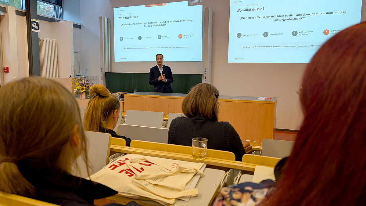Ein Mann steht in einem Hörsaal vor Menschen und spricht. Hinter ihm ist eine Präsentation zu erkennen. Unscharf im Vordergrund befinden sich die Hörsaalbänke, teilweise gefüllt.