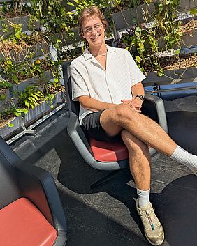 Ein junger Mann mit mittellangen braunen Haaren und Brille sitzt auf einem schwarzen Ledersessel mit rotem Sitzkissen. Er trägt eine kurze schwarze Hose und ein lockeres weißes Oberteil sowie Tennissocken und sportliche Schuhe. Im Hintergrund befinden sich verschiedene grüne Pflanzen.