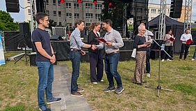 Zu sehen ist eine Preisübergabesituation. Drei Personen werden geehrt, es werden Hände geschüttelt und eine Urkunde überreicht.