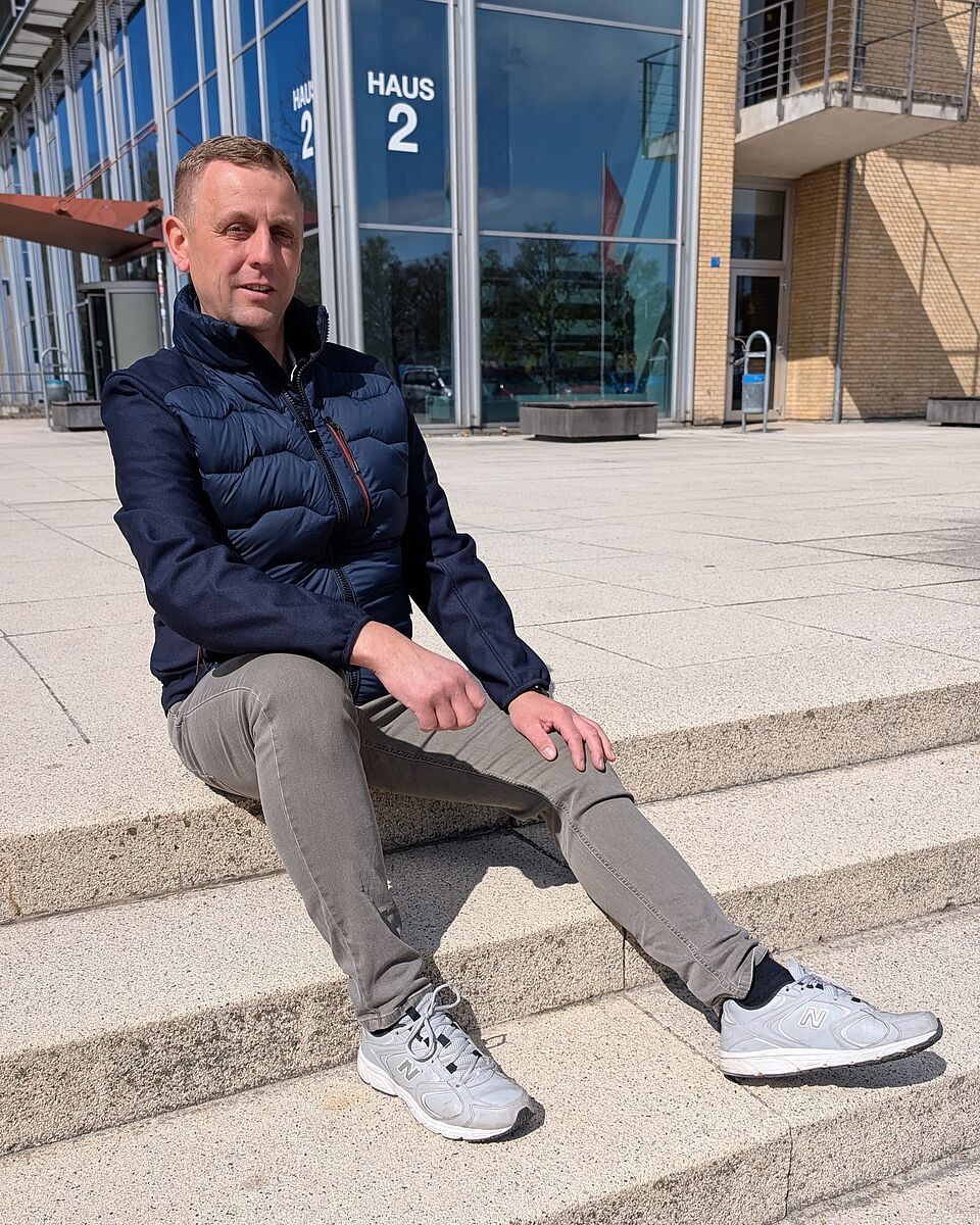 Ein Mann in dunkelblauer Jacke sitzt auf den Treppen vor Haus 2 der Hochschule Neubrandenburg. Er schaut freundlich in die Kamera.