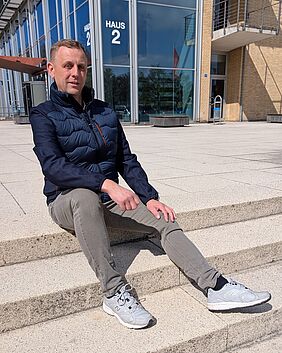Ein Mann in dunkelblauer Jacke sitzt auf den Treppen vor Haus 2 der Hochschule Neubrandenburg. Er schaut freundlich in die Kamera.