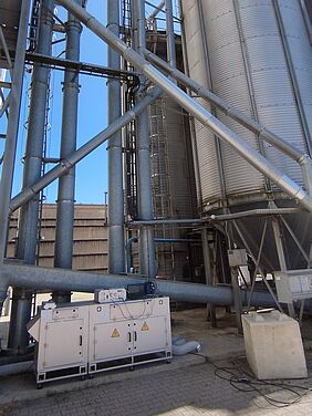 Zu sehen ist ein Silo eines landwirtschaftlichen Betriebs. Davor befindet sich eine Apparatur, optisch vergleichbar mit einer weißen Kommode. Auf und an dem Gerät befinden sich Kabel und gelbe Warnzeichen.