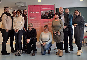Zehn Personen posieren für ein gemeinsames Gruppenfoto vor einer Flipchart, auf der sich ein großes rotes Plakat mit der Aufschrift „MV tut was“ befindet. Zwei Personen knien direkt davor, die anderen haben sich neben dem Plakat versammelt.
