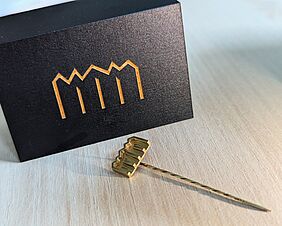 Im Vordergrund liegt eine vergoldete Nadel, die am Kopf das Signet der Hochschule trägt. Im Hintergrund befindet sich eine dunkle Schatulle mit demselben Zeichen.